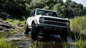 A white 2021 Ford Bronco SUV crosses rocky water with trees in background