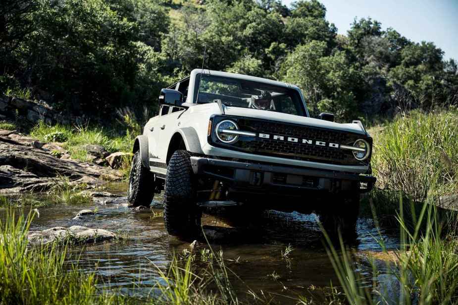 A white 2021 Ford Bronco SUV crosses rocky water with trees in background