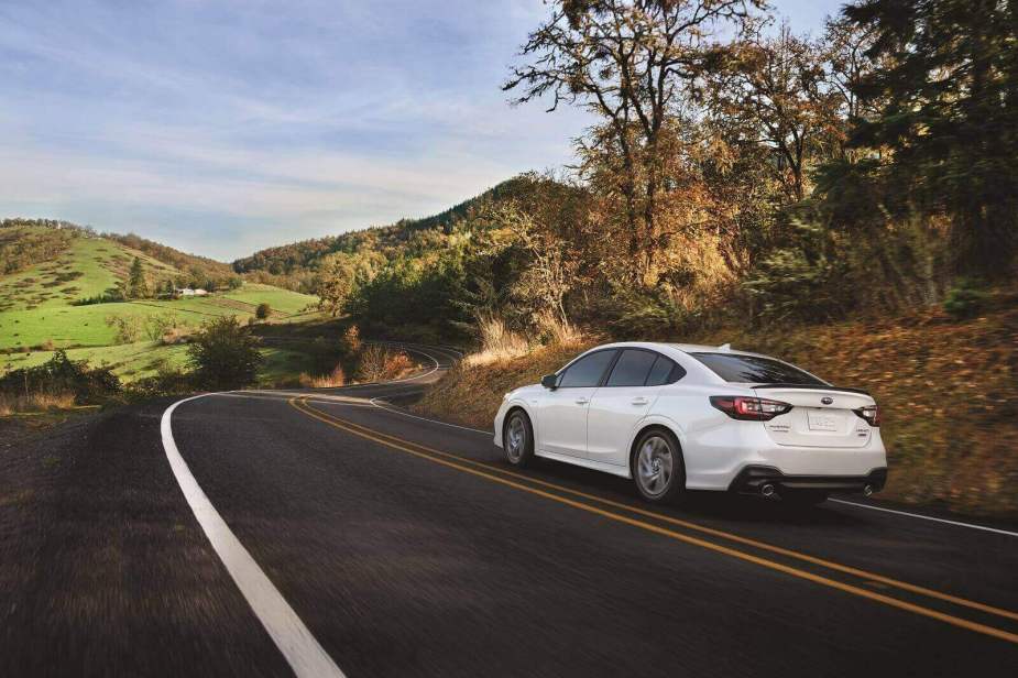 A white 2024 Subaru Legacy drives on a winding hilly road away from the viewer in left rear angle