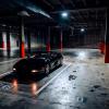 A black Ferrari F355 sits under overhead lights in a parking garage.