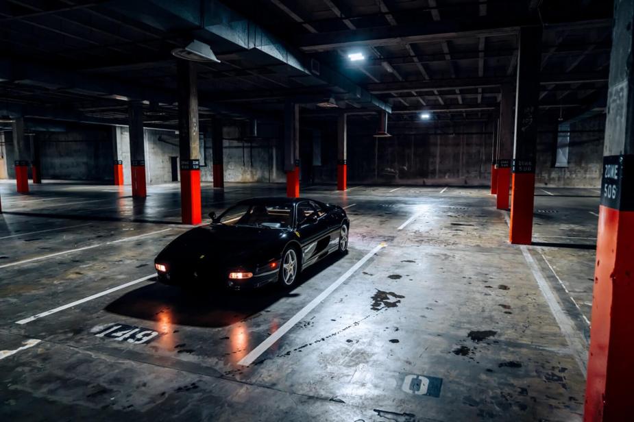 A black Ferrari F355 sits under overhead lights in a parking garage.