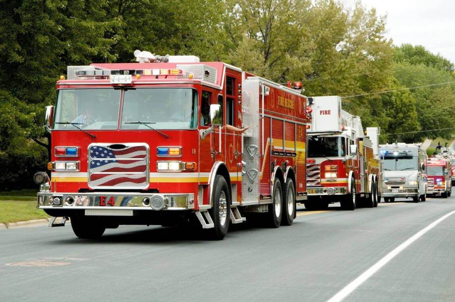 A parade of fire trucks cruise past a gas station on a busy street.