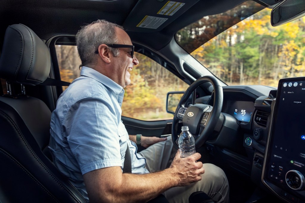 A man smiles widely while sitting in a Ford F-150 Lightning using BlueCruise hands-free driving tech man holding water bottle in right hand resting other hand on leg