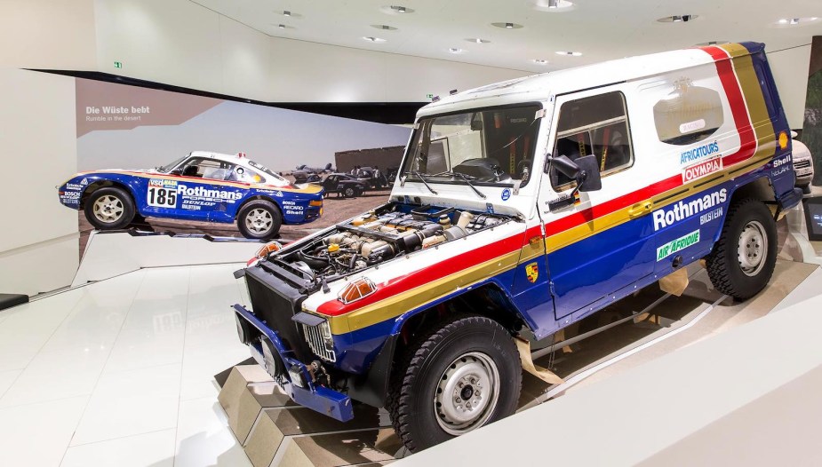A blue and white Mercedes G-Class SUV and Porsche race car parked in a museum.