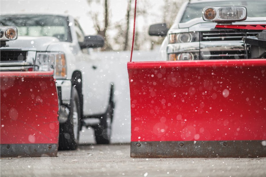 Red plows on a pair of white pickup trucks parked in the snow.