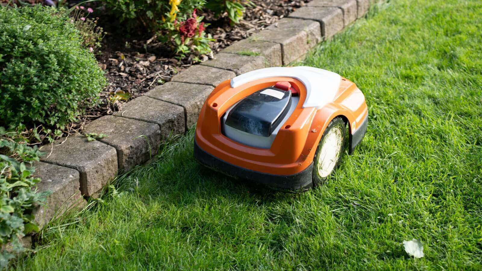 A robot lawn mower mows a lawn next to a little wall.