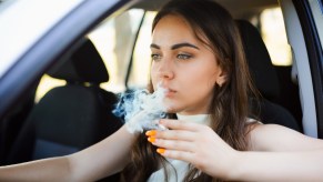 Young woman holds a cigarette while driving, and exhales through the open window of her car