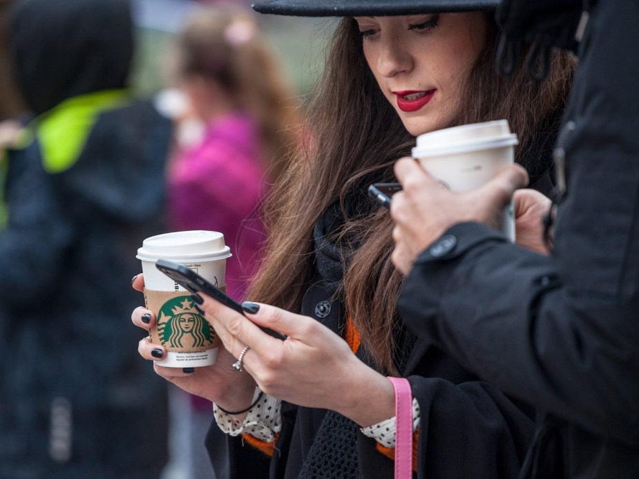 Two young women scroll on their phones while drinking their starbucks coffee