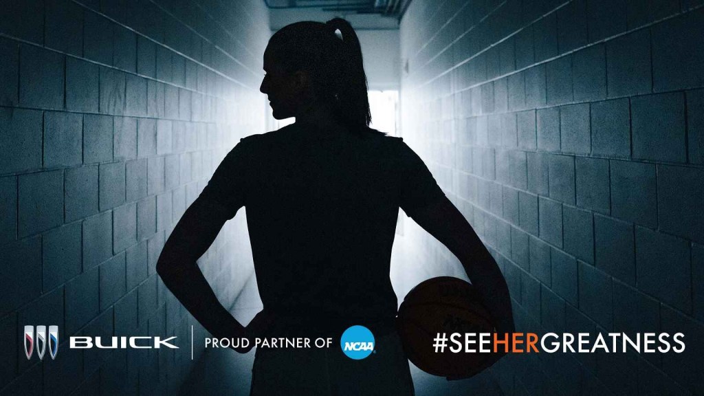 Caitlin Clark shown in dark outline in the tunnel walk to basketball court head turned to her right holding basketball on left hip