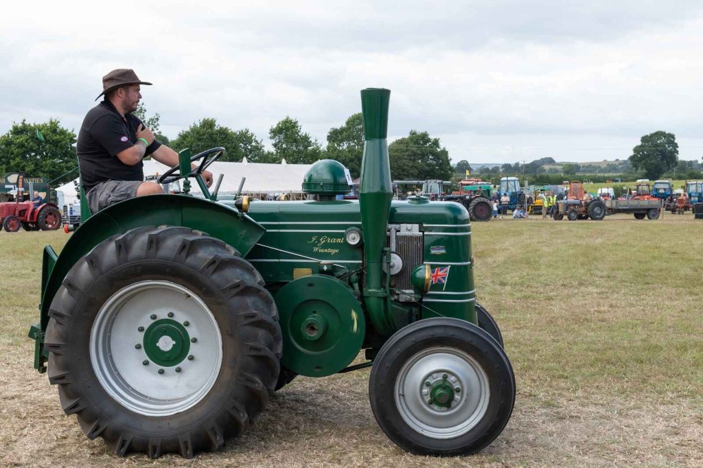 A green vintage Field Marshall tractor being driven by a man wearing a hat facing right profile view