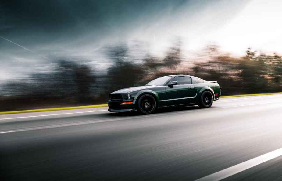 A dark colored Ford Mustang shown driving fast on a blurred road in left front profile view