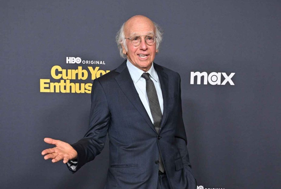 Larry David stands in front of an HBO Max Curb Your Enthusiasm banner in a dark suit and tie