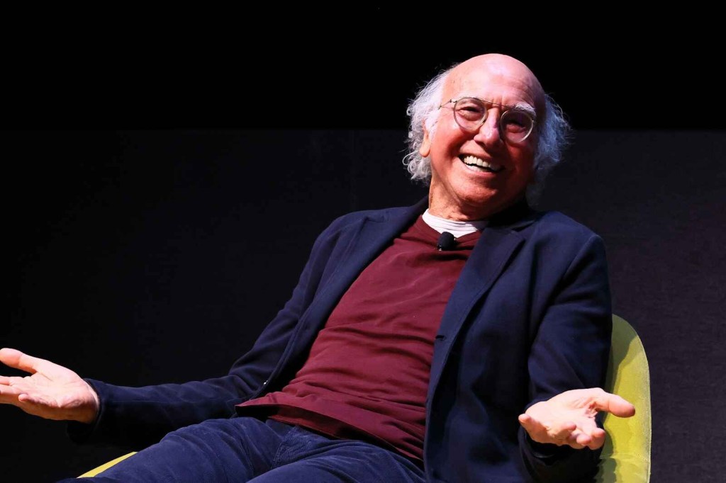 Larry David sitting in a yellow chair under stage lighting in close torso view smiling with open arms palms up