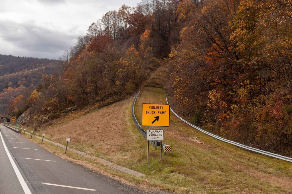 A runaway truck ramp is shown center frame on hilly autumn-wooded freeway