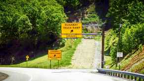 A yellow runaway truck ramp sign with a "gravel" sign posted under it in front of the ramp green trees grassy hill
