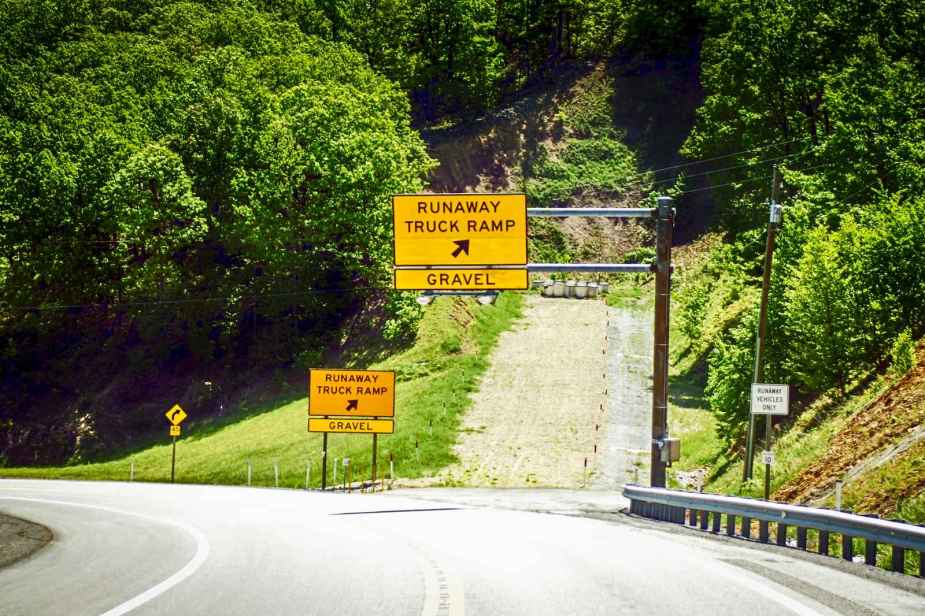 A yellow runaway truck ramp sign with a "gravel" sign posted under it in front of the ramp green trees grassy hill