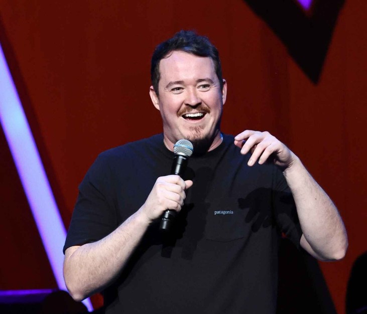 Comedian Shane Gillis on stage wearing a black t-shirt talking into a microphone smiling