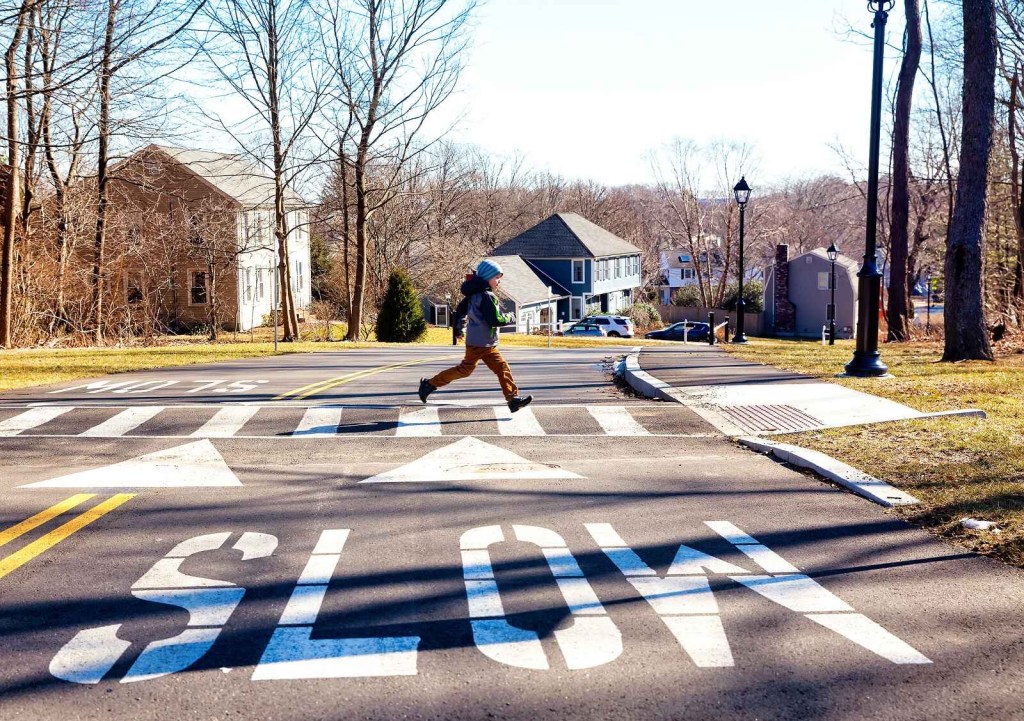 A child runs across a street on a crosswalk with "SLOW" spelled in white paint on the pavement in front of viewer