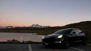 A black Tesla Model 3 parked in left front angle view with headlines glowing at deep dusk in front of water and snow capped mountain