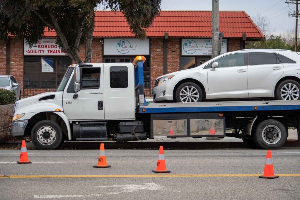 A rollback tow truck with a silver SUV on the bed in left profile view with cones in the foreground