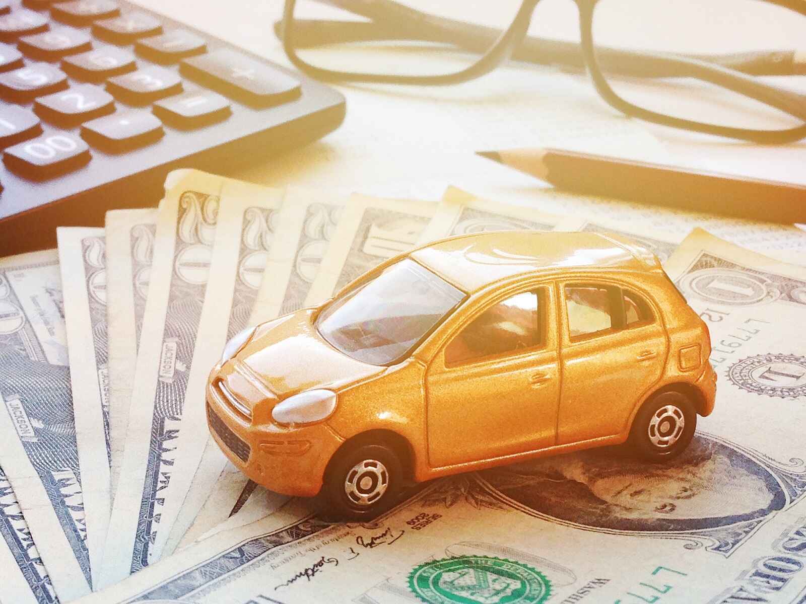A gold toy car sits on a stack of American paper money with a handheld calculator, pencil, and reading glasses in the backgound