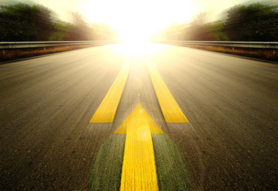A bright sun at the end of a blurred road with high contrast orange stripes and painted arrow pointed down the road