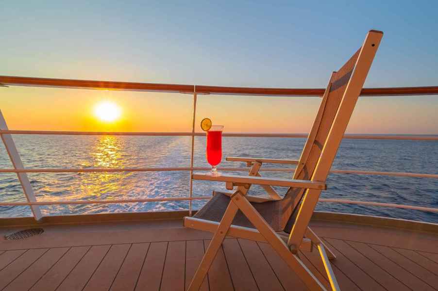 A wooden folding chair with pink cocktail on arm sits on a cruise ship deck at sunset