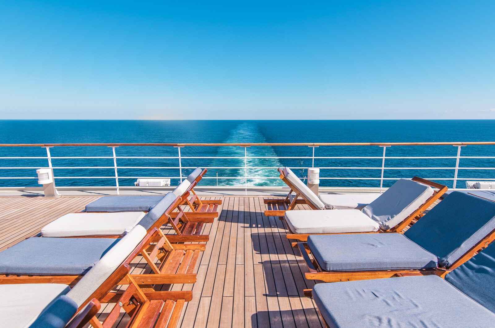 A cruise ship deck with rows of cushioned wood loungers with ship wake trailing center background clear blue sky