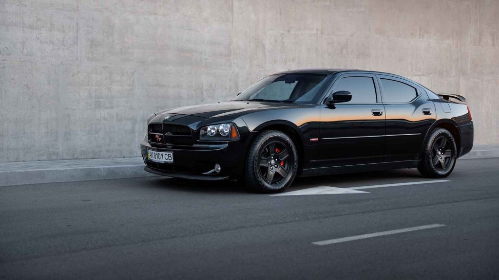 A black Dodge Charger sedan driving on a paved road next to a concrete wall facing left in profile view