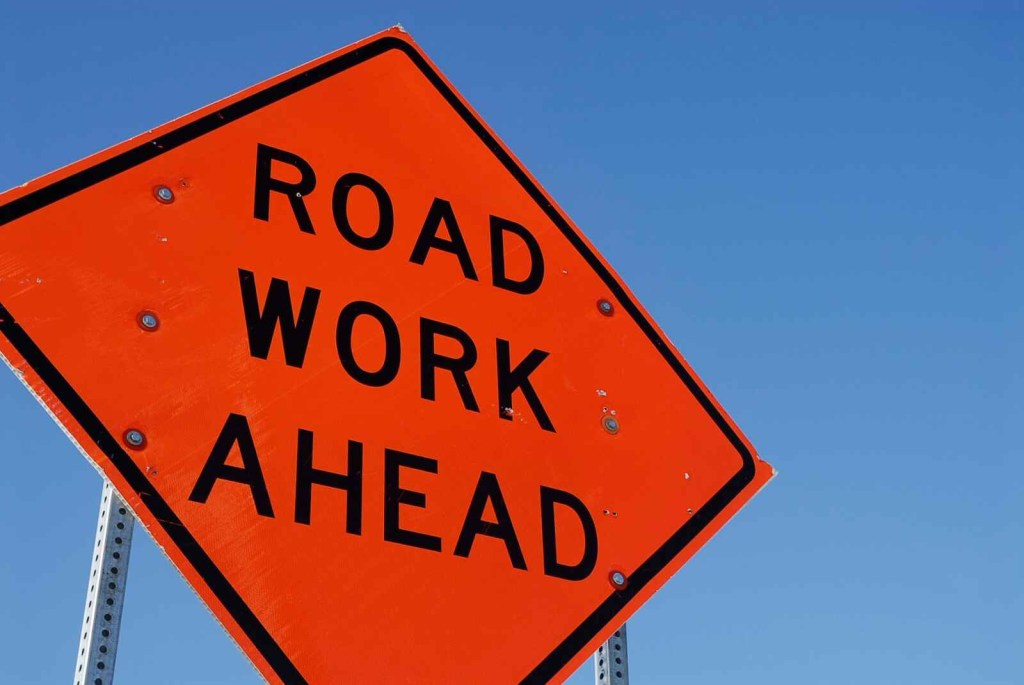 A dark orange sign with "ROAD WORK AHEAD" in black lettering with blue sky background