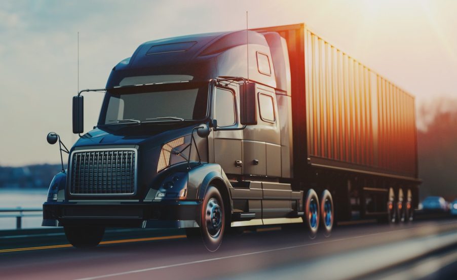 A semi-truck in close left front angle view driving on a highway with blurry sun in right upper corner