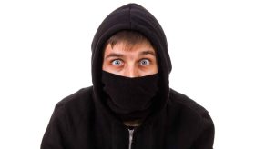 A depiction of a wide-eyed thief wearing black mask covering nose and mouth and black hoodie sweatshirt blue wide eyes looking straight at viewer