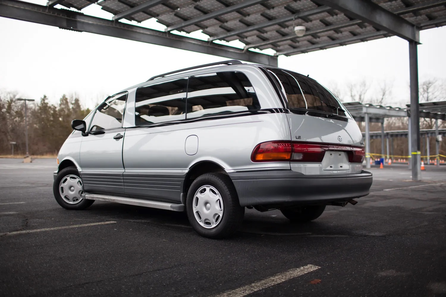A silver Toyota Previa van released in 1994 is parked in a lot at left rear angle overcast weather perfect 90s road trips cars