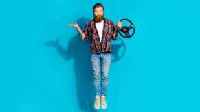 A man with a long beard dark red hair jumping in front of a blue screen holding a steering wheel prop in one hand with other arm in a palm-up shrug position