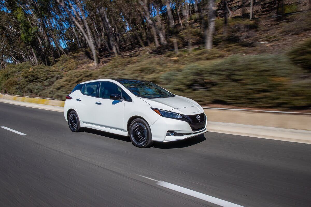 A white Nissan LEAF drives by a tree line. 