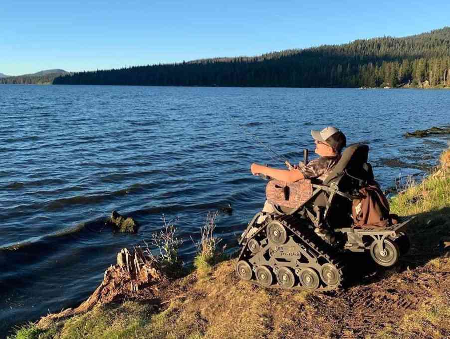 Man fishes from a lakeshore while sitting in an all-terrain trackchair.