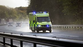 A neon yellow checkered British ambulance races down a UK highway in the rain.