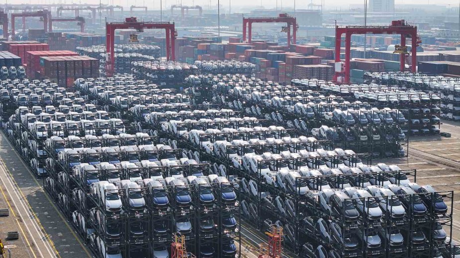 Countless Chinese EVs stacked on racks on a dock before export on a ship in the background.