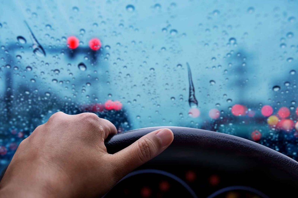 A view of a human hand holding a car steering wheel in front of windshield in very rainy weather close view