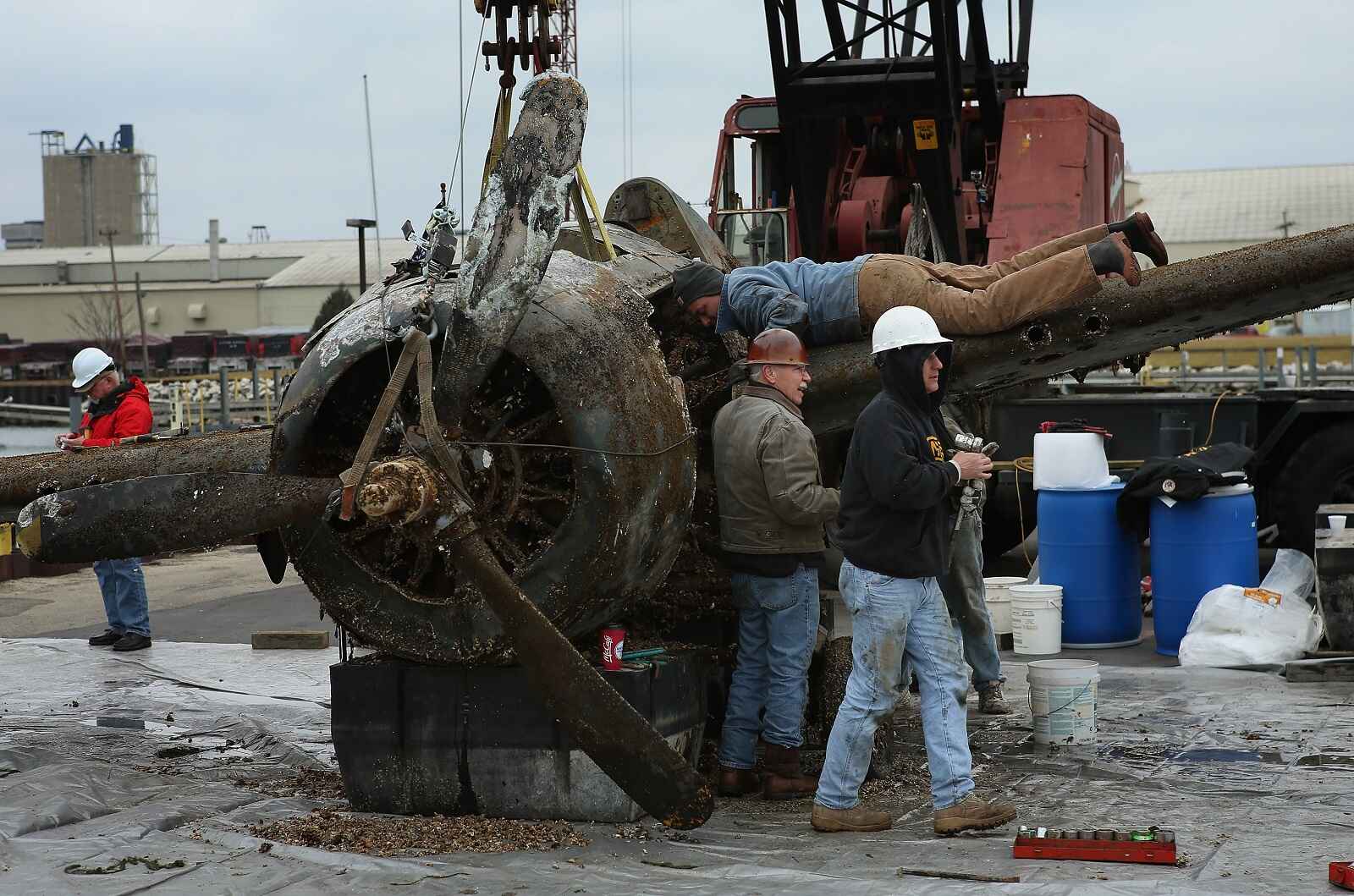 A WWII-era fighter plane craned out of Lake Michigan with a crew surrounding it and man laying on left wing