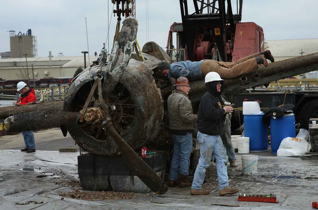 A WWII-era fighter plane craned out of Lake Michigan with a crew surrounding it and man laying on left wing