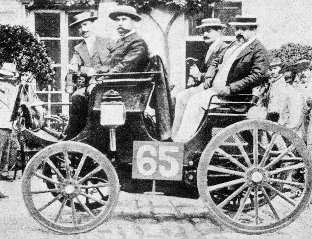 Four men sit in an early race car on the streets of paris