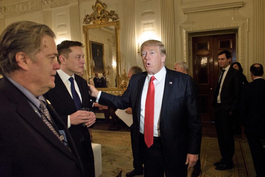 Donald Trump greets Elon Musk in the white house, placing a hand on the Tesla CEO's shoulder, while advisor Steve Bannon looks on.