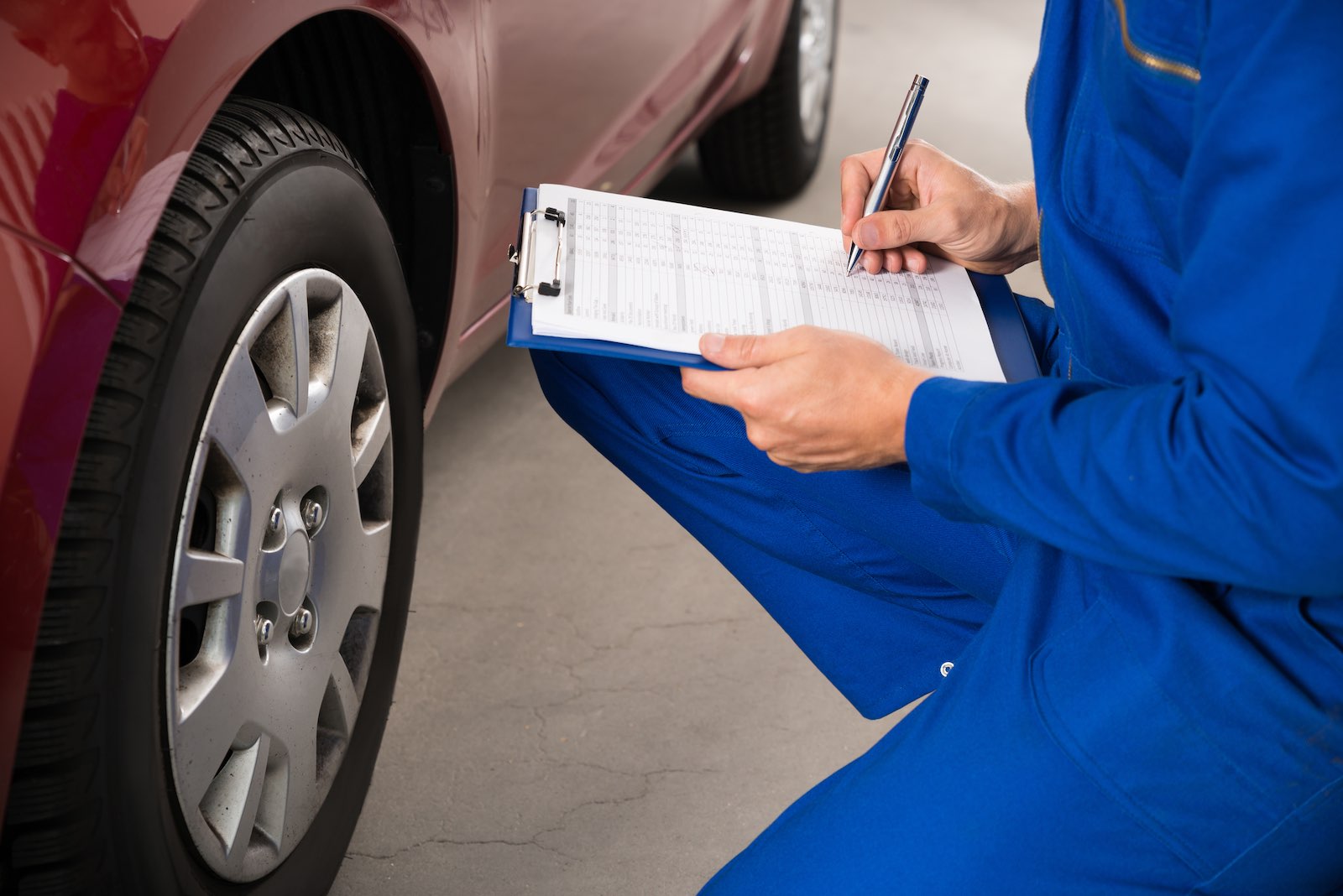 Mechanic in blue coveralls inspects a car, taking notes on a clipboard.