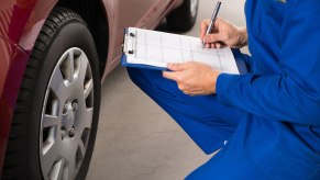 Mechanic in blue coveralls inspects a car, taking notes on a clipboard.