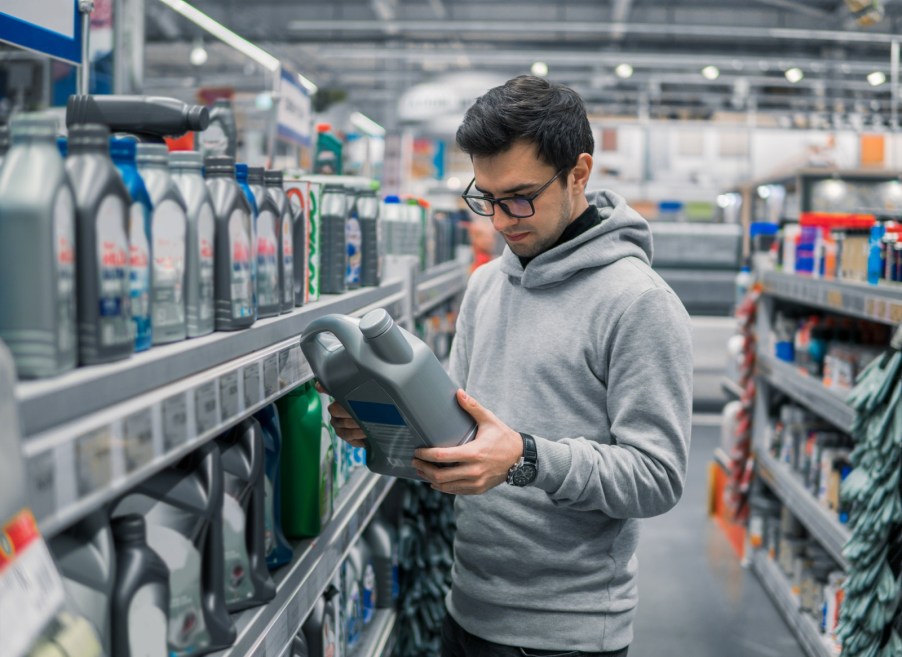 Male in grey hoodie and black glasses holds a bottle of car engine oil reading label in a store shelves full of engine oil options