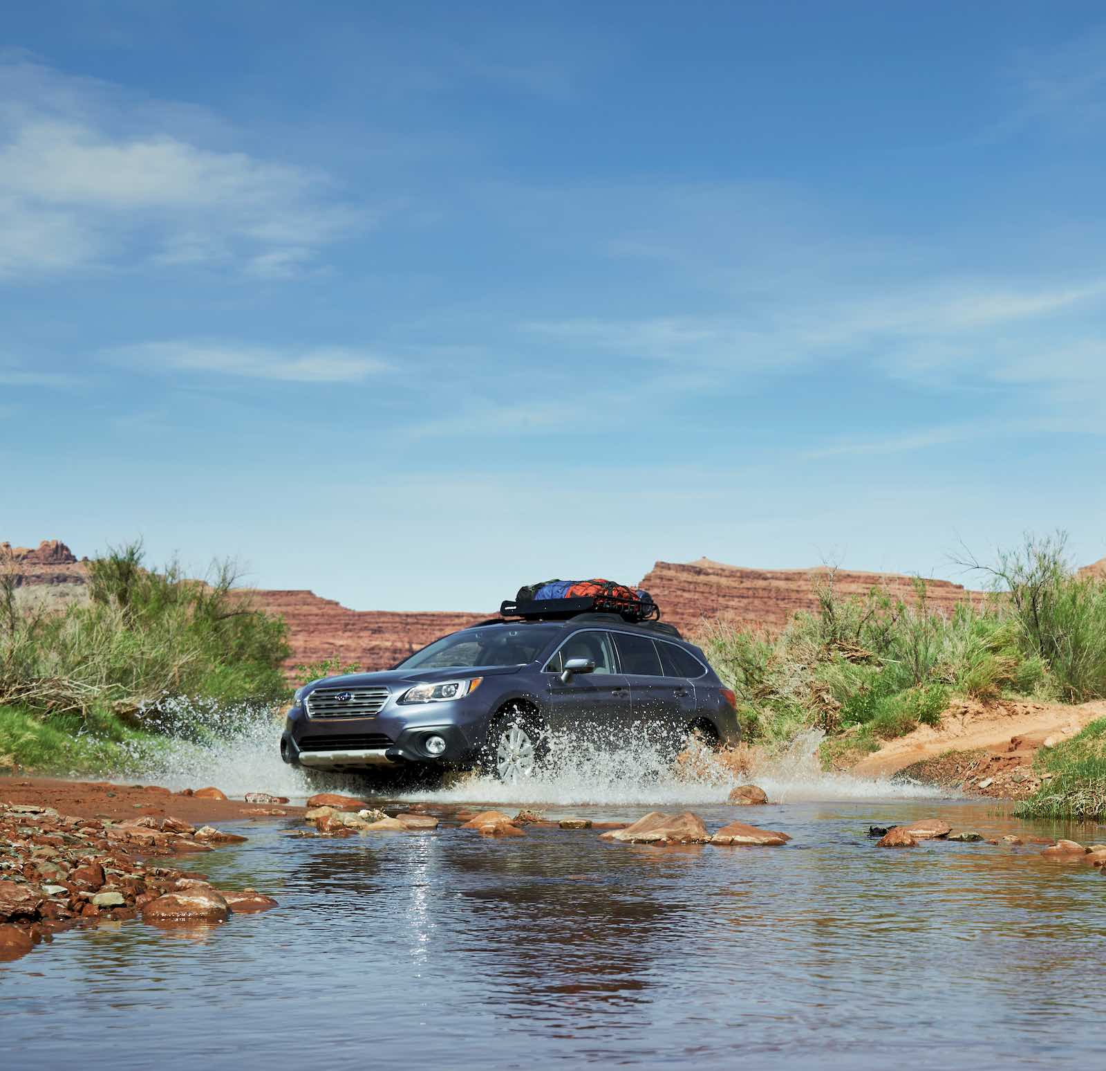 A Subaru Outback drives through a stream, mountains visible in the background.