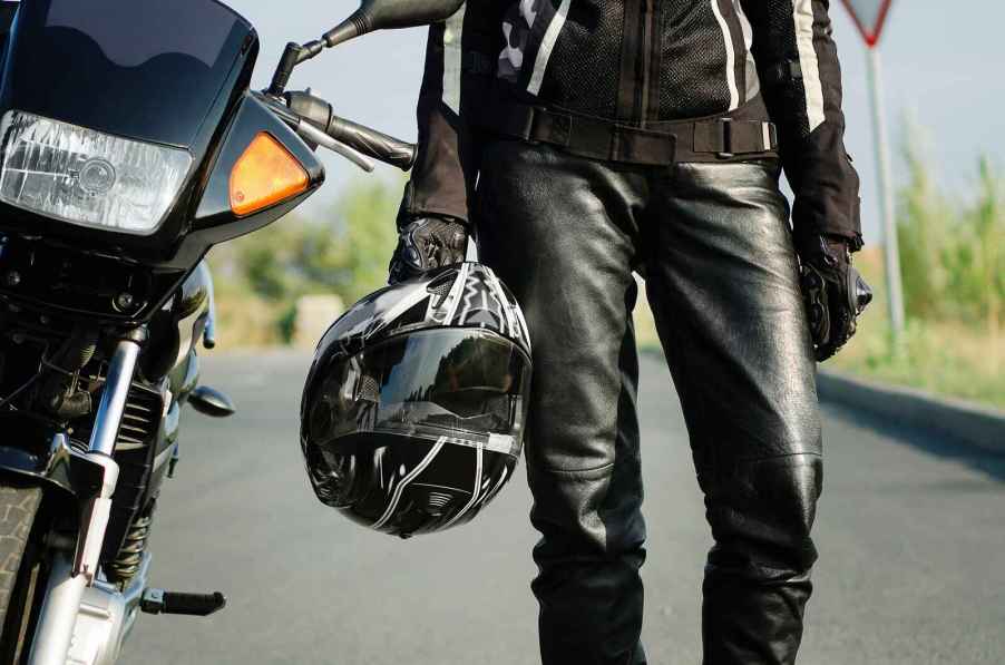 Motorcycle rider wearing leather gear holding a helmet shown from torso to shins