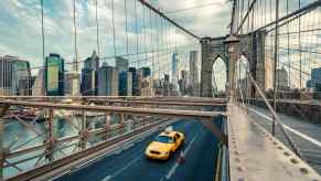 Bright yellow New York City taxi cab crosses the Brooklyn bridge, Manhattan's skyline visible in the background.
