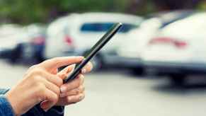 A woman's hands shown holding a cell phone in front of a blurred row of cars parked outside